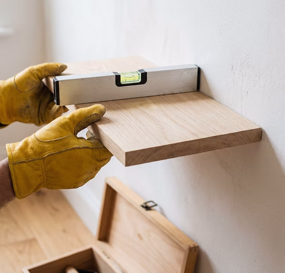 Gloved hands checking a wooden shelf with a spirit level while mounting it on the wall